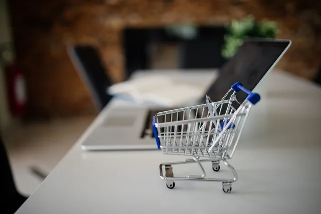 A miniature shopping cart sits prominently on a desk next to a sleek laptop, suggesting the theme of online shopping or e-commerce in a contemporary workspace. Lagkrav på e-handelsplattformar i Sverige